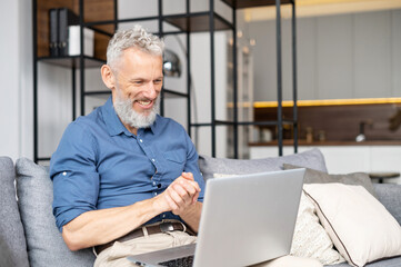 Modern middle-aged bearded man using laptop computer sitting on the couch at home, spending leisure time online, watching movies. Mature male working remotely from home, looks at the screen and smiles