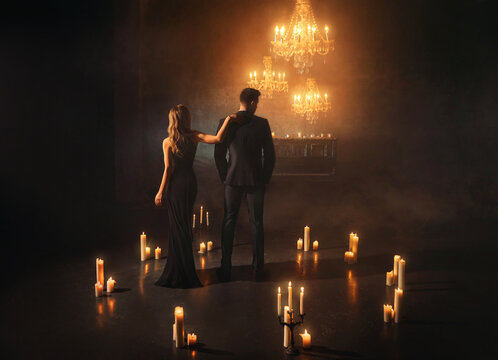 gothic couple stands in dark room woman hugs man in business suit. black elegant dress. ball carnival party, luxurious scenery many candles burning. Fantasy girl vampire queen, guy king back rear view