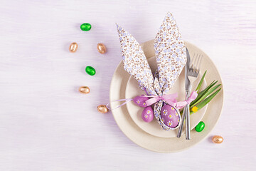 Easter table setting. White plate with a napkin folded in the shape of a rabbit, Easter and chocolate eggs on a pink background. Happy Easter holiday concept. Top view, flat lay