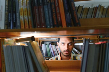 Portrait of cheerful male international Indian student with backpack, learning accessories standing near bookshelves at university library or book store during break between lessons. Education concept