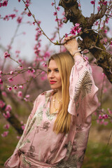 The portrait of young blondie woman in pink silk costume sitting at the branch  enjoying the blossom peach garden 