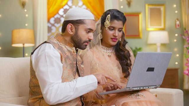 Happy smiling muslim couple busy using laptop during festival celebration while sitting on sofa at home - concept of technology, family boding and e-commerce