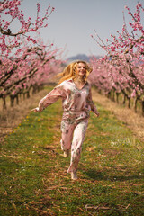 The young blondie woman having fun jumping smiling in pink silk costume into blossom peach garden 