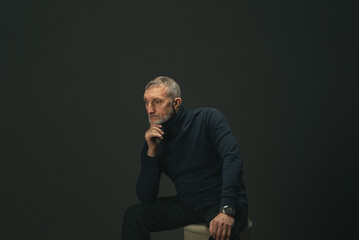 an elderly man with a beard is sitting on a pouf in the studio on a dark background, he was thinking about something