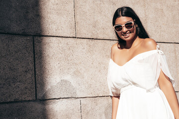 Young beautiful smiling hipster woman in white dress. Sexy carefree model posing on the street background at sunset. Positive female outdoors at warm sunny day. Cheerful and happy. In sunglasses