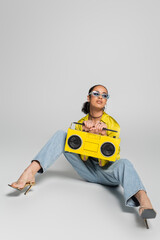 full length of brunette african american woman in blue sunglasses posing with yellow boombox on grey.