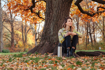 Young woman sitting near tree in autumn park spend time outdoors, drinking coffee or tea. Pretty lady in yellow coat resting in fall nature. Concept of relax and leisure activity. Copy ad text space