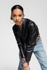 fashionable african american woman in trendy cropped jacket and blue jeans looking at camera isolated on grey.