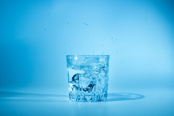Ice cubes fall into a glass of water on a blue background. Refreshing drinking water