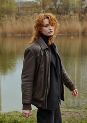 a beautiful red-haired woman walks in the park near the lake in a leather jacket, enjoying a pleasant day