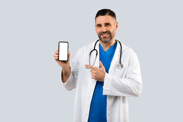 Cheerful middle aged general practitioner man in uniform pointing at cellphone blank screen, grey...