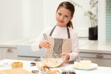 Obraz na płótnie Canvas Cheerful caucasian little girl in apron make cookie dough with whisk in kitchen interior, copy space