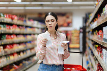 Portrait of young amazement Caucasian woman by high prices in supermarket. Shocked millennial holds...