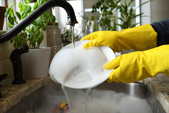 Washing rinsing dishes in kitchen sink by hand wearing yellow rubber gloves.