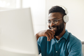 Happy black man in headphones working on computer, watching webinar, freelancing, taking part in online conference