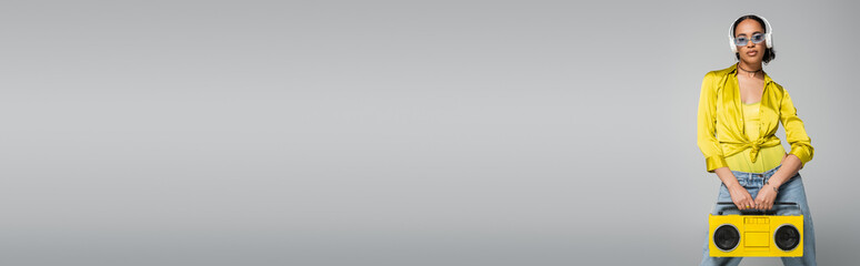 stylish african american woman listening music in wireless headphones and holding boombox on grey, banner.