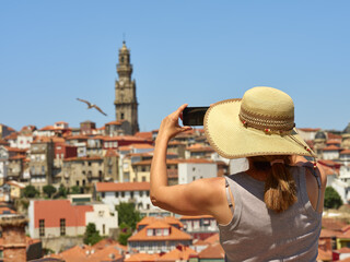 Fototapeta premium Woman wearing a straw hat to protect herself from the sun photographing the panorama of the historic city of Porto, Portugal. In the background the Torre de los Clérigos
