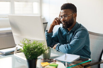 Thoughtful black male ceo working on computer in office, sitting at workplace and thinking about business project
