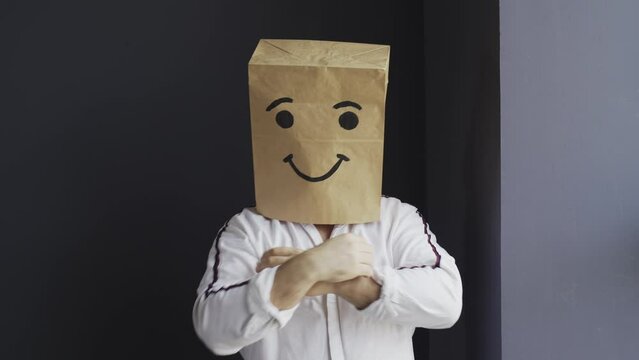 A man in a white shirt with a paper bag on his head, with a painted smiley face, stands against the wall, overwhelmed with feelings, shows a class gesture, with his hand.
