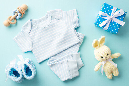 Baby shower concept. Top view photo of infant clothes blue shirt pants knitted booties present box with bow bunny toy and wooden rattle on isolated pastel blue background