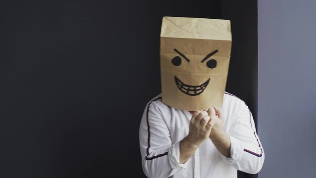 An angry man in a white shirt with a bag on his head, with an evil smiley face drawn, stands in the corner against the wall, laughs, waves his fists. Emotions and gestures.