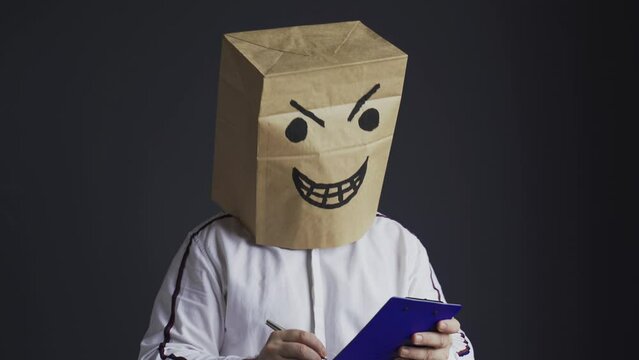 An angry man in a white shirt with a bag on his head, with a drawn angry smiley face, holds a tablet in his hands and crosses out from the list of candidates for the award.
