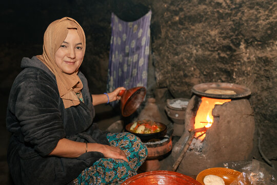 Moroccan berber woman in typical old kitchen with wood oven looking at camera and showing food in tajin