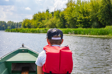 A young boy in a baseball cap and a red life jacket on a green boat is sailing among the trees on a sunny day. Summer. - 586648797