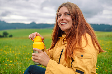 Pretty young girl traveling sitting in mountains drinking tea from camping thermos enjoying trip, tourist student is relaxing in nature. High quality photo