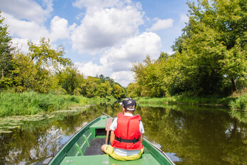 A young boy in a baseball cap and a red life jacket on a green boat is sailing among the trees on a sunny day. Summer. - 586649729