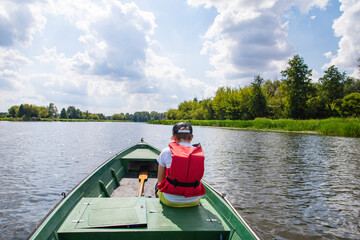 A young boy in a baseball cap and a red life jacket on a green boat is sailing among the trees on a sunny day. Summer. - 586650112