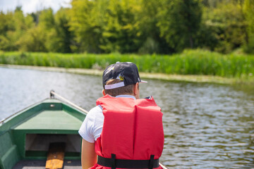 A young boy in a baseball cap and a red life jacket on a green boat is sailing among the trees on a sunny day. Summer.