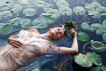 Young sexy unhappy crazy longing woman lady redhead Ophelia with curly red hair lies with water lilies, eyes closed in white dress dying in the water in the lake, in lily pond