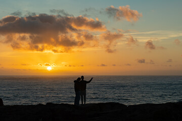 Obraz premium A couple standing on the cliff and watching the sun setting down over the sea