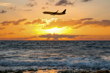 Naklejka na ściany i meble Plane flying over sea during sunset. Sun shining through clouds