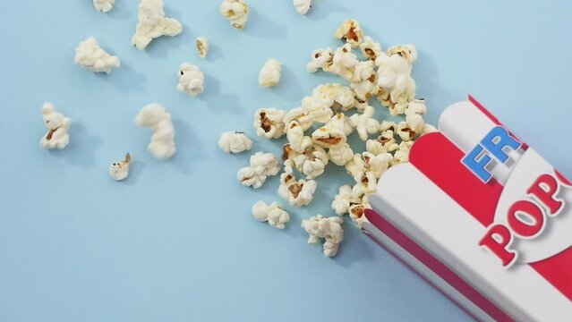 From right bottom corner falls striped paper box full of fresh popcorn and it spreads around blue surface, classical container, movie time, entertainment, fast food. Close up, 4K Ultra HD.