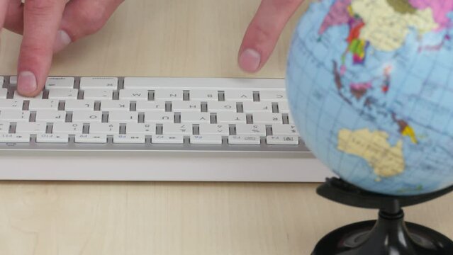 The fingers are typing on the white keyboard on the table next to the globe. The concept of communication on the Internet, booking a trip online, worldwide business.