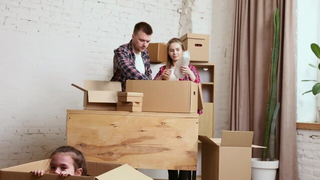 Young happy family, man, woman and little daughter moving into new flat, apartment. Opening many cardboard boxes with stuffs. Playful kid. Concept of moving houses, real estate, family, new life