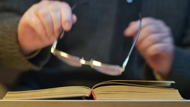An unrecognizable man in a dark sweater puts on glasses and reads an old book at a table in the library.