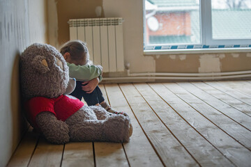 a nine-year-old girl is upset, she is sitting in a room without repairs on the floor, hugging her knees and crying, next to a large soft toy bear, copy space