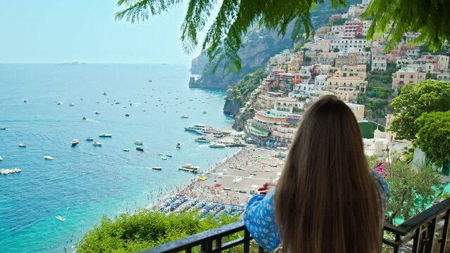 A female tourist exploring the cliffside of Positano. A young woman looking from above over the beautiful colorful houses and boats with the turquoise beach in Positano, Amalfi Coast.