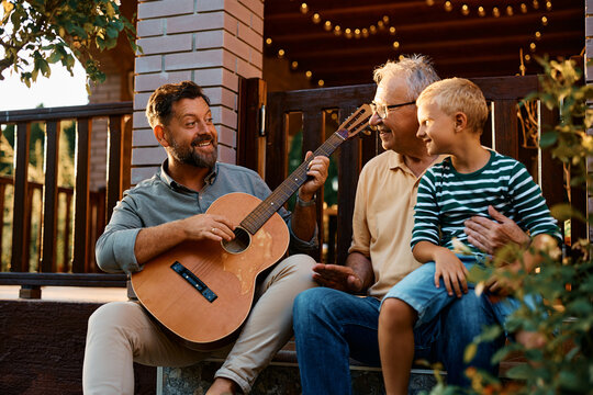 Happy man plays acoustic guitar to his son and senior father in backyard.
