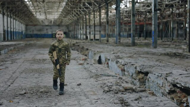 Little Boy in Military Uniform Walking in Destroyed Plant Holding a Toy Gun on the Ruins Background. Patriot Soldier Kid. Boy Playing in War