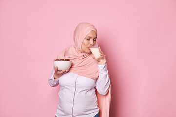 Lovely Muslim pregnant woman with head covered in pink hijab, holding a bow full of fresh dates and taking sip of delicious vegan milk, isolated on pink background. Healthy breakfast during pregnancy