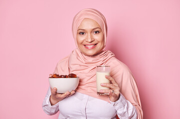 Close-up portrait of a cheerful Muslim woman in pink hijab smiling at camera, holding a glass with milk and a bowl with fresh dates, ideal breakfast during Ramadan fasting, isolated on pink background