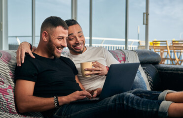 Male couple smiling while using a laptop sitting on the couch at home.