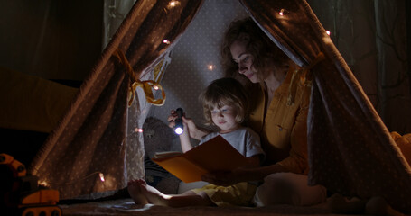 Happy caucasian mom, cute baby boy and funny dog spending time together at home, reading a book, using a flashlight in a cozy tent - happy family 