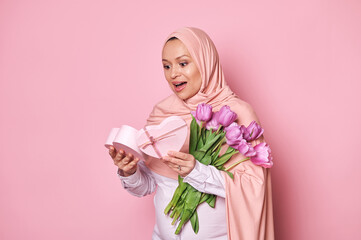 Amazed Muslim woman in pink hijab, expressing happiness, receiving a cute present in heart shaped gift box, posing with a bouquet of tulips on isolated background. International Women's, Mother's Day