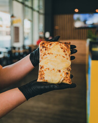 Un french tacos gratiné à la raclette, présenté dans les mains d'une jeune femme portant des gants noirs