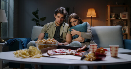 Young caucasian couple spending time in front of tv together, being scared by a horror movie and eating unhealthy food 